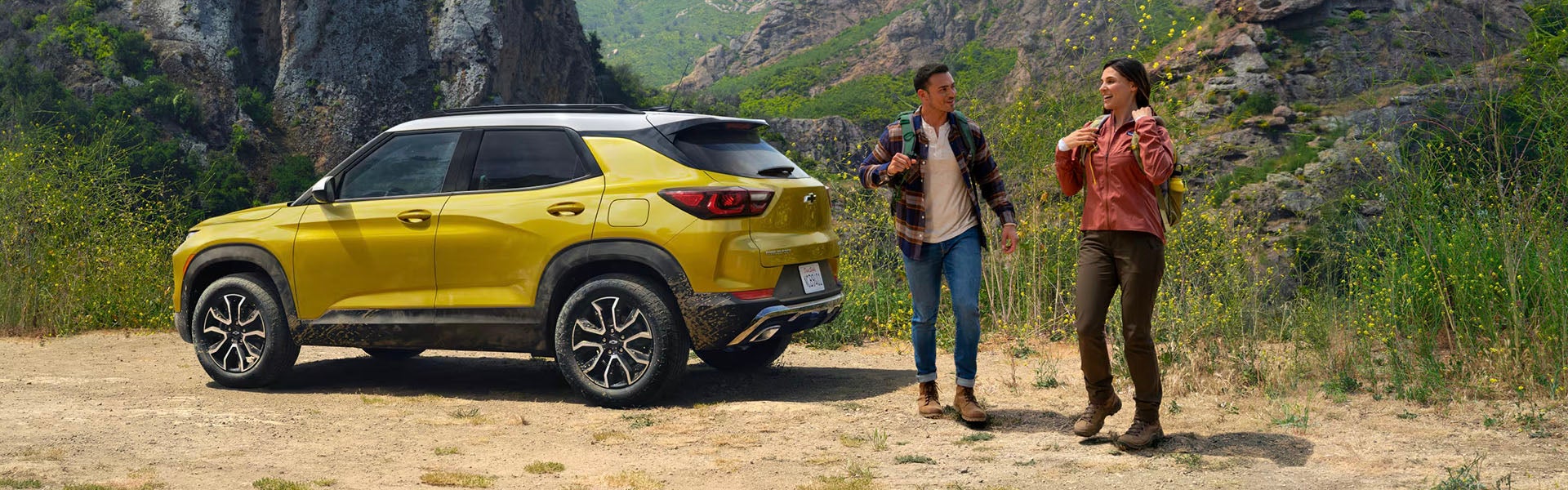 A yellow Chevrolet Trailblazer parked near two hikers in a mountainous landscape