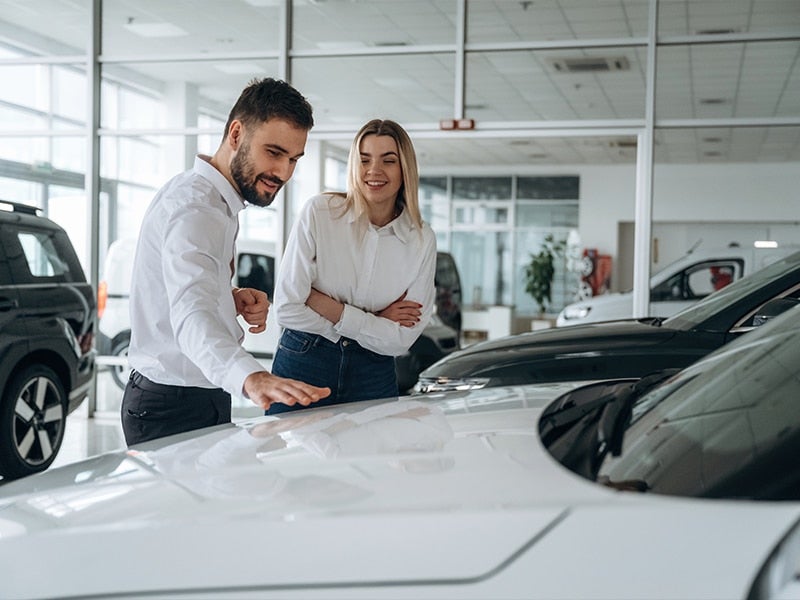 Man in a car dealership talking to a smiling woman while pointing at the hood of a white car.