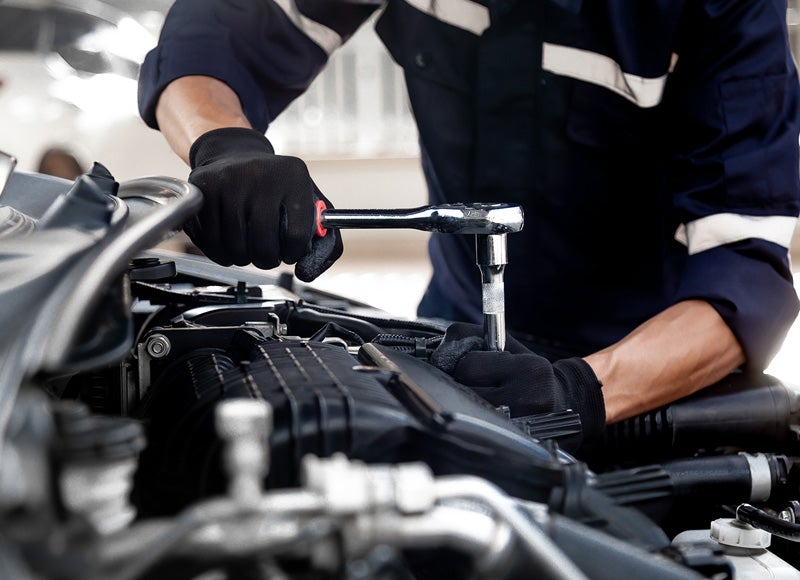A mechanic in gloves uses a ratchet wrench to work on a car engine under the open hood.